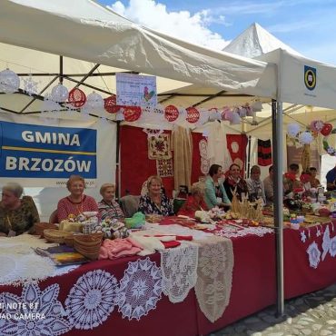 Grupa osób siedzi przy stoliku, na którym leżą różnorodności do zakupu (hafty, koszyki itp.