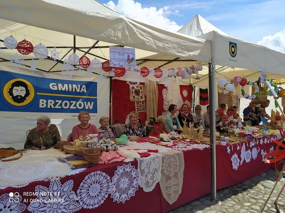 Grupa osób siedzi przy stoliku, na którym leżą różnorodności do zakupu (hafty, koszyki itp.