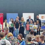 Grupa dzieci i okien osoba starsza stoją na scenie otrzymują dyplom i puchar.