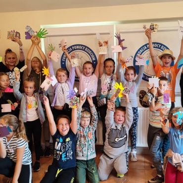 Grupa dzieci pozuje do zdjęcia w jasnej sali z rękoma uniesionymi do góry i machającymi.