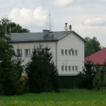 Budynek z jasną elewacją z nowym połozonym dachem koloru szarego, przed nim rosną drzewa.