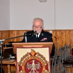 Starszy mężczyzna ubrany w strażacki mundur galowy przemawia z mównicy.