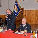 Dwaj mężczyźni ubrani w mundury Galowe siędzący przy stole z czerwonym obrusem z czego jeden z nich przemawia do mikrofonu