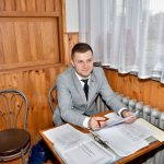 Mężczyzna ubrany w szary garnitur z czarnym krawatem siedzi przy stole.