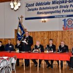 Druhowie i druhny siedzą przy stole z czerwonym obrusem ubrani w mundury galowe. Jeden z nich przemawia.