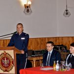 Pan policjant ubrany w mundur galowy wstaje i przemawia przez mikrofon z mównicy.