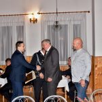 Mężczyźni ubrani w czarny garnitur i mundur galowy składają podziękowania mężczyźnie ubranego w granatowy garnitur. Dołącza do nich mężczyzna ubrany w białą koszulę