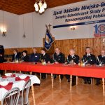 Mężczyźni i kobieta siedzący za stołem z czerwonym obrusem na tle baneru "Zjazd oddziau Miejsko-Gminnego związku OSP RP"