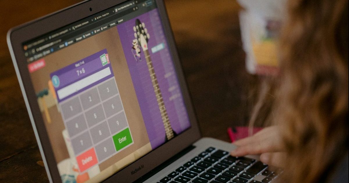 Młoda dziewczyna z kręconymi brązowymi włosami siedzi przy laptopie ucząc się tabliczki mnożenia