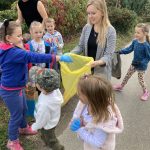 Grupa dzieci w towarzystwie dwóch pań nauczycielek zbiera śmieci do worków. W tle krzewy oraz drzewa.