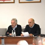 Dwóch mężczyzn w średnim wieku elegancko ubranych, siedzących przy biurku.