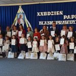 Grupa dzieci i dorosłych (kobiet i mężczyzn) ubranych odświętnie pozuje do zdjęcia w jasnej sali. Za nimi granatowa tablica przyozdobiona napisami Dzień Papieski.