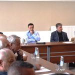 Grupa mężczyzn ubranych elegancko siedzi w jasnej sali. Przed nimi leżą dokumenty i woda do picia.