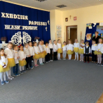 Grupa dzieci ubranych odświętnie dziewczynki zólte spodniczki i białe koszulki. chłopcy granatowe spodnie i białe koszule mówią wierszyki w jasnej sali.