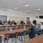 Grupa osób ubranych elegancko siedzi w jasnej, dużej sali obrad. Przed nimi leżą dokumenty i woda do picia.