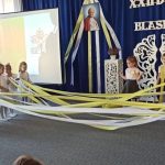 Grupa dzieci dziewczynek i chłopców tanczy z długimi wstążakami biało zółtymi na jasnej sali.