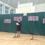 Starsza kobieta ubrana w czarną sukienkę przemawia do mikrofonu. Za nią ustawiona jest zielona tablica.