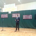 Młody mężczyzna ubrany w ciemny garnitu stoi w jasnej sali i przemawia do mikrofonu. Za nim postaiowna jest zielona tablica ze zdjęciami.