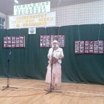 Kobieta ubrana w jasną długą suknie z kapeluszem na głowie przemawia do mikrofonu na sali gimnastycznej.