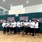 Grupa kobiet ubranych na biało czarno śpiewają do mikrofonu na jasnej sali.