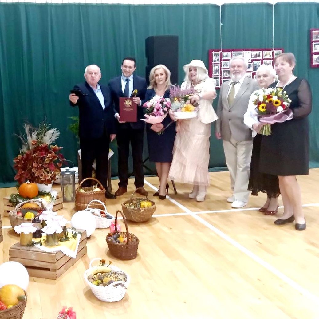 Grupa osób ubranych elegancko stoi w jasnej sali gimnstycznej. Przed nimi ustawiona jest dekoracja składająca sie z drewnianych skrzynek, koszy i warzyw.