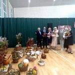 Grupa osób ubranych elegancko stoi w jasnej sali gimnstycznej. Przed nimi ustawiona jest dekoracja składająca sie z drewnianych skrzynek, koszy i warzyw.