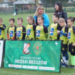 Grupa młodych chłopaków ubranych w zielone stroje piłkarskie stoi na murawie, za nimi dwie kobiety jedna młodsza o czarnych włosach, druga o blond włosach, przed nimi baner.