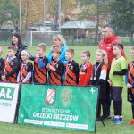 Grupa młodych chłopaków ubranych w stroje piłkarskie stoi na murawie, za nimi dwie kobiety jedna młodsza o czarnych włosach, druga o blond włosach, przed nimi baner.