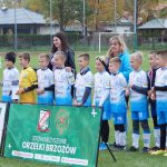 Grupa młodych chłopaków ubranych w stroje piłkarskie stoi na murawie, za nimi dwie kobiety jedna młodsza o czarnych włosach, druga o blond włosach, przed nimi baner.