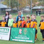 Grupa młodych chłopaków ubranych w stroje piłkarskie stoi na murawie, za nimi dwie kobiety jedna młodsza o czarnych włosach, druga o blond włosach, przed nimi baner.