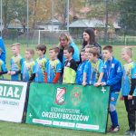 Grupa młodych chłopaków ubranych w stroje piłkarskie stoi na murawie, za nimi dwie kobiety jedna młodsza o czarnych włosach, druga o blond włosach, przed nimi baner.