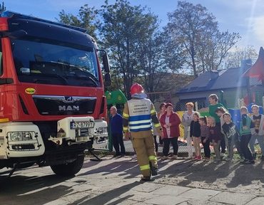 Grupa dzieci stoi przed czerwonym wozem strażackim, przed nim strazak w mundurze rozmawia z grupą kilkunastu dzieci,
