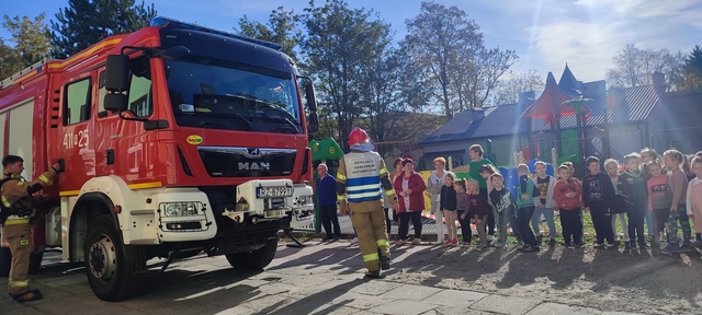 Grupa dzieci stoi przed czerwonym wozem strażackim, przed nim strazak w mundurze rozmawia z grupą kilkunastu dzieci,