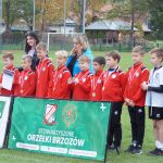 Grupa młodych chłopaków ubranych w stroje piłkarskie stoi na murawie, za nimi dwie kobiety jedna młodsza o czarnych włosach, druga o blond włosach, przed nimi baner.