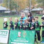 Grupa młodych chłopaków ubranych w stroje piłkarskie stoi na murawie, za nimi dwie kobiety jedna młodsza o czarnych włosach, druga o blond włosach, mężczyzna w zielonym sportowym stroju przed nimi baner.