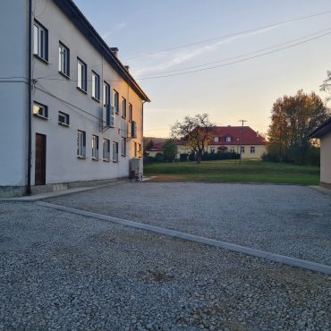 Jasny budenk z elewacją, obok plac wyłożony kamieniem, na przeciwko budynek jasny z czerownym dachem szkoły.