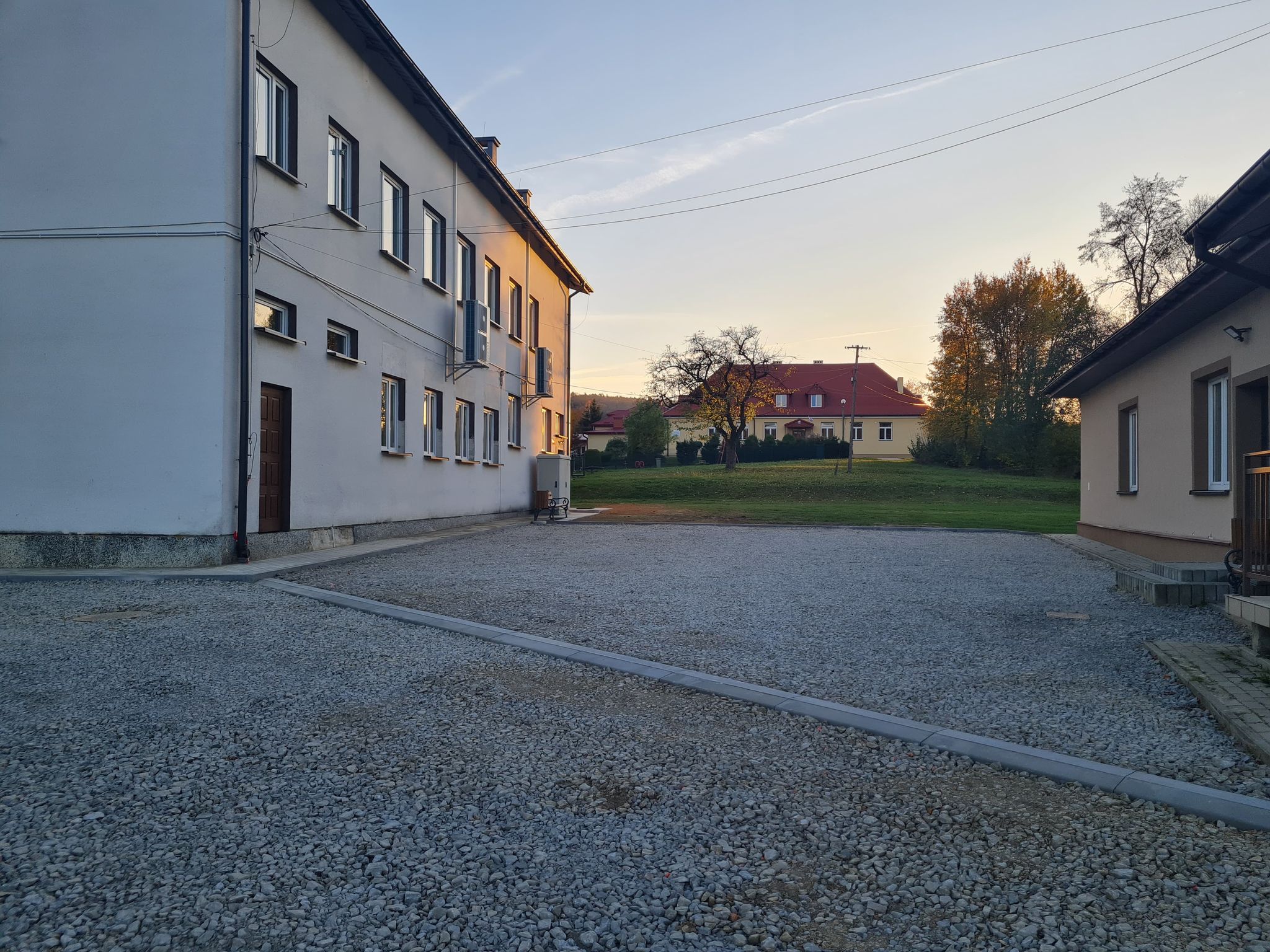 Jasny budenk z elewacją, obok plac wyłożony kamieniem, na przeciwko budynek jasny z czerownym dachem szkoły.