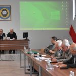 Grupa osób ubranych elegancko siedzi w jasnej, dużej sali obrad. Przed nimi leżą dokumenty i woda do picia. Na ścianie jest wyświetlana prezentacja multimedialna.