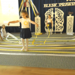 Grupa dzieci dziewczynek i chłopców tańczy z biało żółtymi wstążakmi na jsanej sali. Za nimi tablica granatowa.