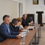 Trzech mężczyzn i kobieta siedzą przy biurku. Przed nimi znajdują się dokumenty i woda do picia. Wszyscy są odświętnie ubrani.