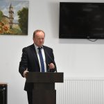 Ubrany w garnitur mężczyzna w średnim wieku, przemawia na sali za pomocą mikrofonu.