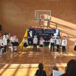 Grupa odświętnie ubranych dzieci w sali gimnastycznej. Są oglądani przez publiczność złożoną z dzieci i starszych.