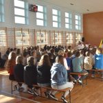 Siedzący na ławkach dzieci i doroślii, znajdujący się w sali gimnastycznej.