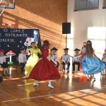 Grupa dzieci ubranych w odświętne stroje. Przed nimi trójka tańczących dziewczynek w sukniach balowych.