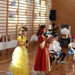 Dwie dziewczynki wystepujace z mikrofonami na sali gimnastycznej. Ubrane są w suknie balowe. Z tyłu siedzą odświętnie ubrane dzieci oraz dorośli.