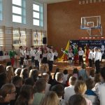 Duża grupa dorosłych i dzieci siedzących na sali gimnastycznej obserwujący występ artystyczny dzieci szkolnych.