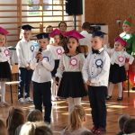 Grupa odświętnie ubranych dzieci występujących w sali gimnastycznej.