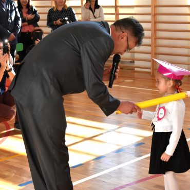Mężczyzna w średnim wieku pasuje dużym ozdobnym ołówkiem dzieczynkę ubraną w biały odświętny strój.