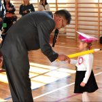 Mężczyzna w średnim wieku pasuje dużym ozdobnym ołówkiem dzieczynkę ubraną w biały odświętny strój.