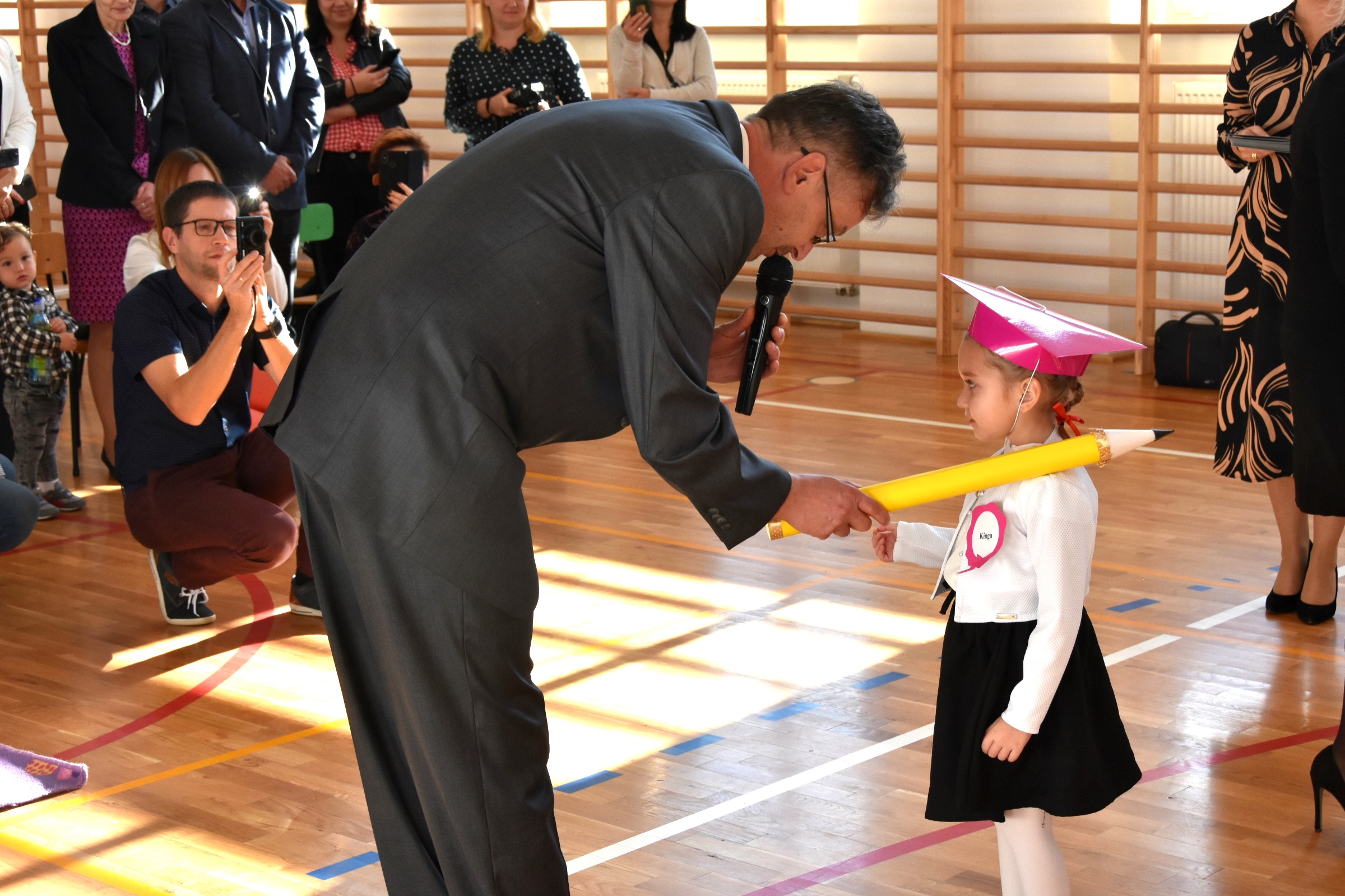 Mężczyzna w średnim wieku pasuje dużym ozdobnym ołówkiem dzieczynkę ubraną w biały odświętny strój.